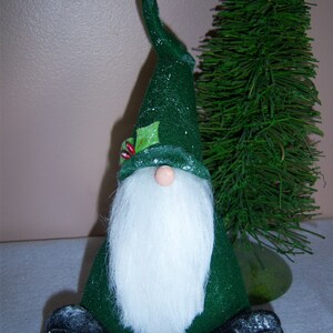 May include: A green felt gnome with a white beard and a white pom-pom on the top of its hat. The gnome is sitting on a white surface.