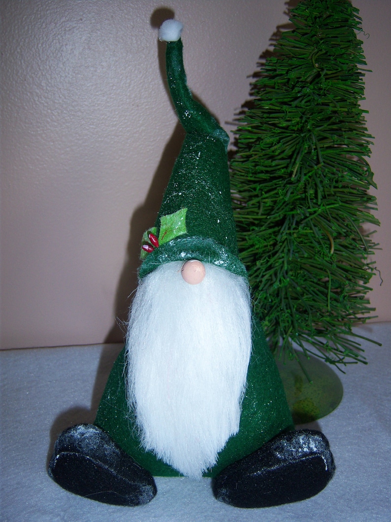 May include: A green felt gnome with a white beard and a white pom-pom on the top of its hat. The gnome is sitting on a white surface.