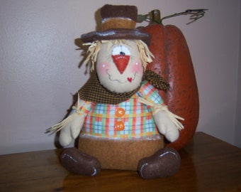 May include: A handmade scarecrow doll with a brown hat, plaid shirt, and straw accents. The scarecrow is sitting next to a large orange pumpkin.