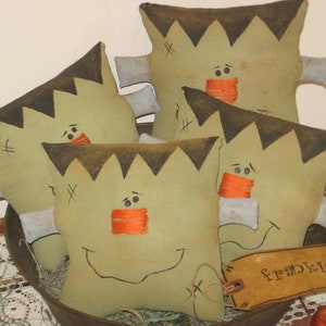 May include: Four green and brown fabric Frankenstein pillows with stitched-on eyes, mouths, and noses. The pillows are arranged in a stack with the top pillow labeled "Frankie".