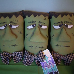 May include: Three green fabric pillows with stitched-on facial features resembling a monster. Each pillow has a black and white checkered bow tie.