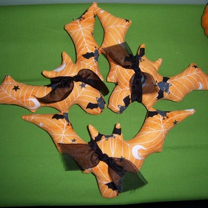 May include: Five orange fabric bats with black spiderwebs and bats printed on them. Each bat has a black ribbon and a sheer black ribbon tied around it. The bats are arranged in a star shape.