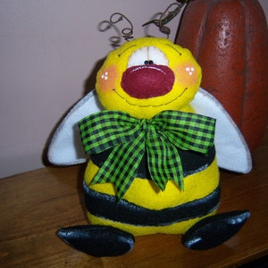 May include: A yellow and black bee-shaped plush toy with a green and black checkered bow tie. The bee has a white felt wing on each side and a red felt nose.