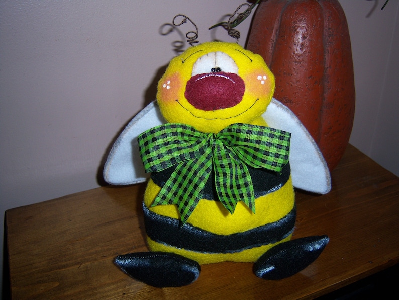 Pode incluir: Um brinquedo de pel&uacute;cia em forma de abelha amarelo e preto com um la&ccedil;o xadrez verde e preto. A abelha tem uma asa de feltro branco de cada lado e um nariz de feltro vermelho.