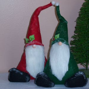 May include: Two plush gnomes, one red and one green, with white beards and green holly accents. They are standing upright with their arms raised.
