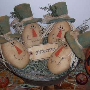 May include: Four handmade scarecrow figures with green hats and brown burlap bodies are arranged in a metal bowl filled with straw. The scarecrows have painted faces with red noses and stitched smiles. A tag with the word "Scarecrows" is visible in the center of the bowl.