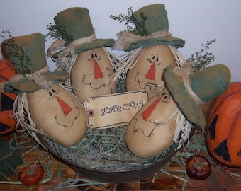 May include: Four handmade scarecrow figures with green hats and brown burlap bodies are arranged in a metal bowl filled with straw. The scarecrows have painted faces with red noses and stitched smiles. A tag with the word "Scarecrows" is visible in the center of the bowl.