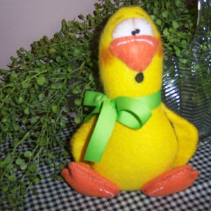 May include: A yellow felt chick with a green bow, orange beak, and black eyes. The chick is sitting on a black and white checkered surface.