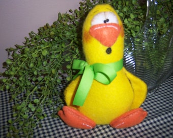May include: A yellow felt chick with a green bow, orange beak, and black eyes. The chick is sitting on a black and white checkered surface.