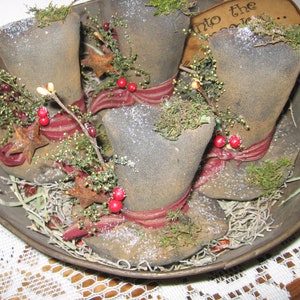 Op de afbeelding: Drie vintage-stijl stoffen hoge hoeden versierd met rode bessen, takjes groen en roestige sterren. De hoeden zijn gerangschikt in een metalen kom met een bruine, rustieke afwerking. De kom is gevuld met gedroogd gras en de tekst "Into the Woods..." is zichtbaar op een klein bord.
