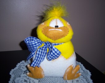 Pode incluir: Decoração de pintinho da Páscoa feita à mão. O pintinho é amarelo com bico e pés castanhos, e um laço xadrez azul e branco. O pintinho está saindo de um ovo branco.