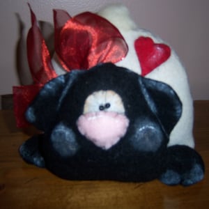 May include: A black plush dog with a pink nose and white eyes, wearing a white hat with a red heart and a red ribbon.