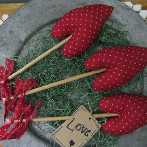 May include: Three red fabric hearts with white polka dots on wooden sticks, arranged on a silver platter with green moss. A brown tag with the word "Love" is in the foreground.