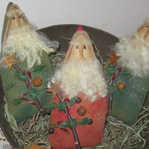 May include: Three handmade felt Christmas ornaments in the shape of Santa Claus. Each ornament is dressed in green and red felt with a white felt beard and a white felt star. The ornaments are sitting in a brown metal bowl filled with straw.