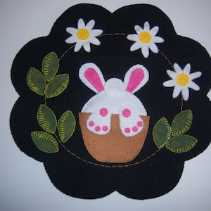 May include: A felt appliqué of a white bunny with pink ears and feet sitting in a brown pot. The bunny is surrounded by green leaves and white flowers with yellow centers.