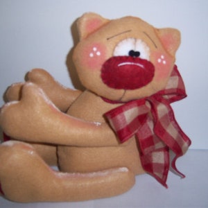 Op de afbeelding: Een bruin pluche kat met een rode neus en een rood-wit geblokte strik. De kat heeft een verdrietige uitdrukking met witte accenten rond de ogen en roze wangen. De armen van de kat zijn om zichzelf heen gewikkeld.