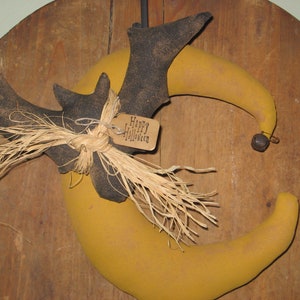 May include: A black bat with straw tied around its wings is hanging from a yellow crescent moon with a brown tag that says "Happy Halloween".