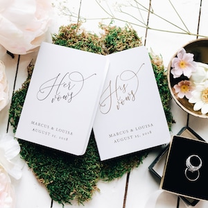 May include: Two white vow books with gold lettering. The books are titled "Her Vows" and "His Vows" with the names Marcus & Louisa and the date August 25, 2018 printed below.