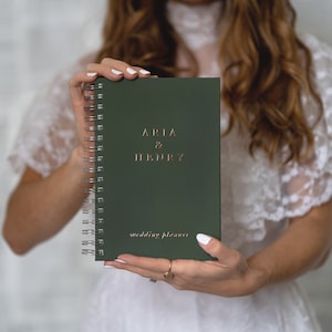 May include: A dark green spiral-bound wedding planner with the names "ARIA & HENRY" in gold lettering. The words "wedding planner" are printed below. The planner is held in front of a white lace background.