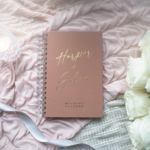 May include: A blush pink wedding planner with gold foil lettering that reads "Harper + Julian" and "Wedding Planner". The planner is on a white and pink textured surface with white roses.