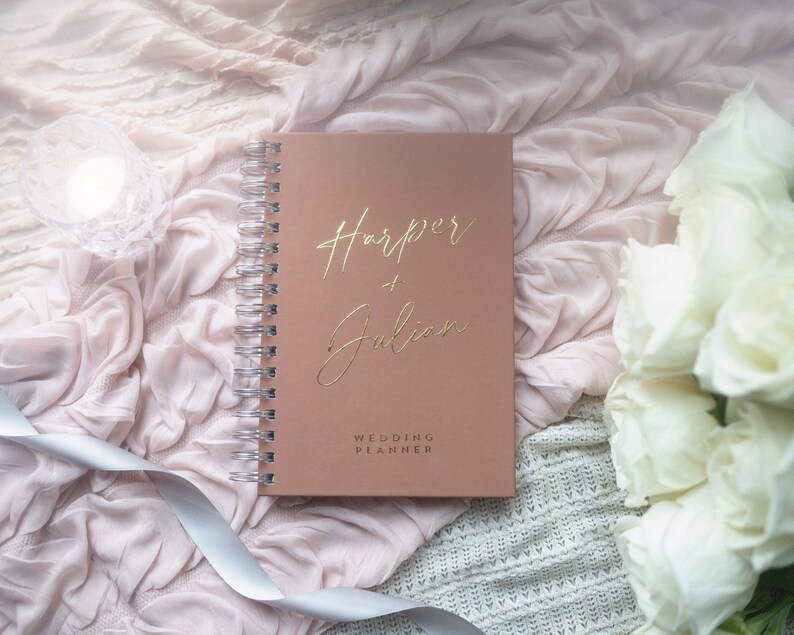 May include: A blush pink wedding planner with gold foil lettering that reads "Harper + Julian" and "Wedding Planner". The planner is on a white and pink textured surface with white roses.