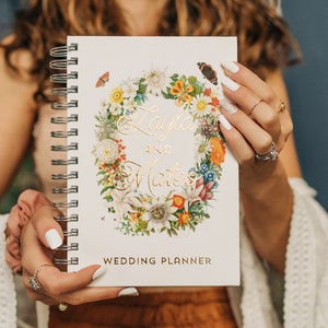 May include: A white wedding planner with a floral wreath design and gold lettering. The planner says "Layla and Mateo" and "Wedding Planner".