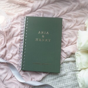 May include: A green wedding planner with silver spiral binding, featuring the names "ARIA & HENRY" in gold lettering. The words "wedding planner" are printed below. The planner rests on a pink fabric with a white ribbon and a bouquet of white roses.