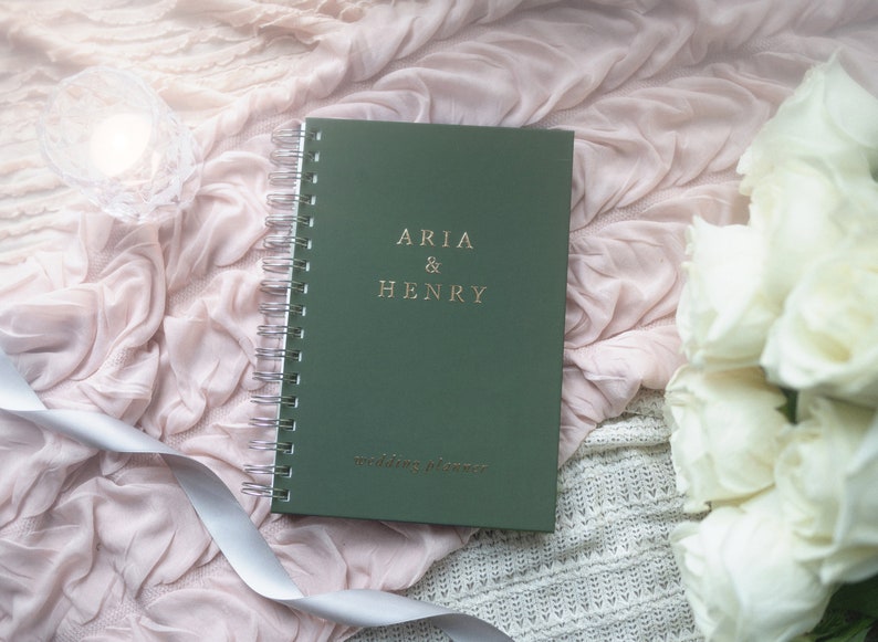 May include: A green wedding planner with silver spiral binding, featuring the names "ARIA & HENRY" in gold lettering. The words "wedding planner" are printed below. The planner rests on a pink fabric with a white ribbon and a bouquet of white roses.