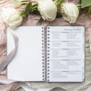May include: An open spiral-bound notebook with a "Wedding Checklist" title, surrounded by white roses, a lit candle in a glass holder, and a satin ribbon. The checklist includes tasks for 12 months to go, 8 months to go, and more.