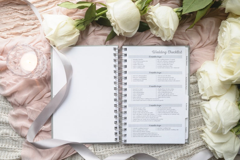 May include: An open spiral-bound notebook with a "Wedding Checklist" title, surrounded by white roses, a lit candle in a glass holder, and a satin ribbon. The checklist includes tasks for 12 months to go, 8 months to go, and more.