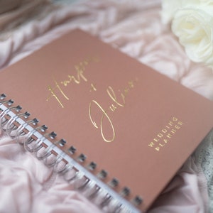 May include: A blush pink wedding planner with gold foil lettering that reads "Happy Ever After" and "Julian". The planner is closed and lying on a white and pink fabric.