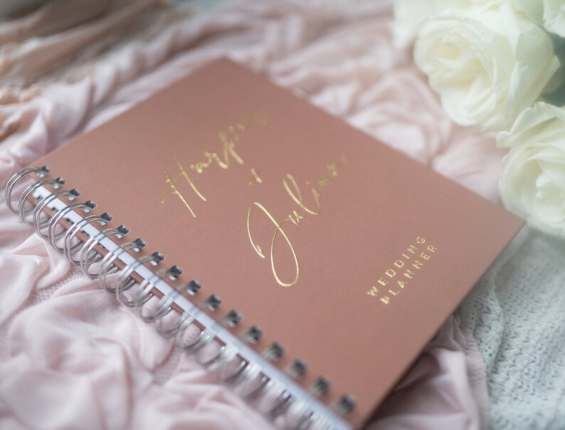 May include: A blush pink wedding planner with gold foil lettering that reads "Happy Ever After" and "Julian". The planner is closed and lying on a white and pink fabric.