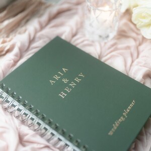 May include: A green wedding planner with spiral binding. The cover features the names "ARIA & HENRY" in gold lettering, along with the words "wedding planner." The planner rests on a pink fabric with flowers and a candle in the background.