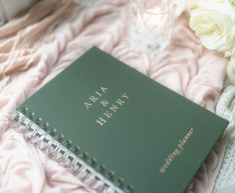 May include: A green wedding planner with spiral binding. The cover features the names "ARIA & HENRY" in gold lettering, along with the words "wedding planner." The planner rests on a pink fabric with flowers and a candle in the background.