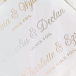May include: A white card with gold, silver, and rose gold foil lettering. The names Olivia & Wyatt, Amelia & Declan, Charlotte & Ezra are written in cursive with ampersands and the type of foil used for each name.