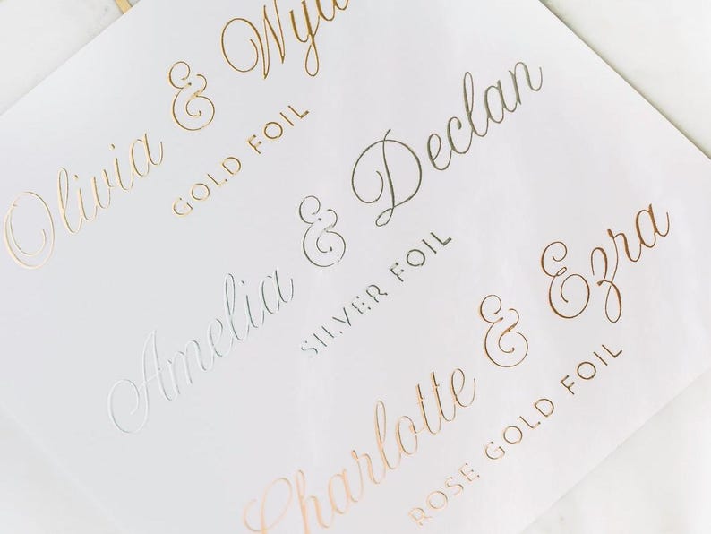 May include: A white card with gold, silver, and rose gold foil lettering. The names Olivia & Wyatt, Amelia & Declan, Charlotte & Ezra are written in cursive with ampersands and the type of foil used for each name.