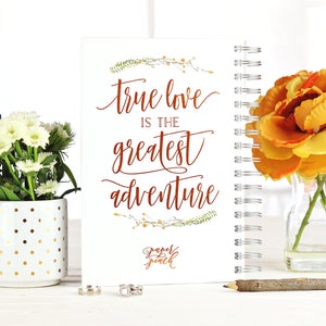 May include: A white spiral-bound notebook with the quote "true love is the greatest adventure" in copper-colored script. The notebook is decorated with floral accents and the "Paper Peach" logo. The image includes flowers and a pencil.