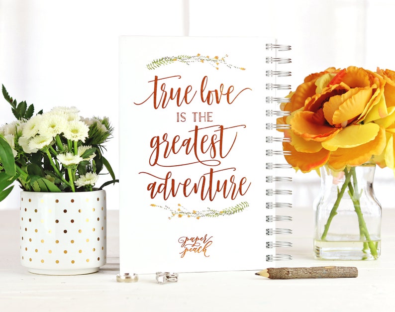 May include: A white spiral-bound notebook with the quote "true love is the greatest adventure" in copper-colored script. The notebook is decorated with floral accents and the "Paper Peach" logo. The image includes flowers and a pencil.