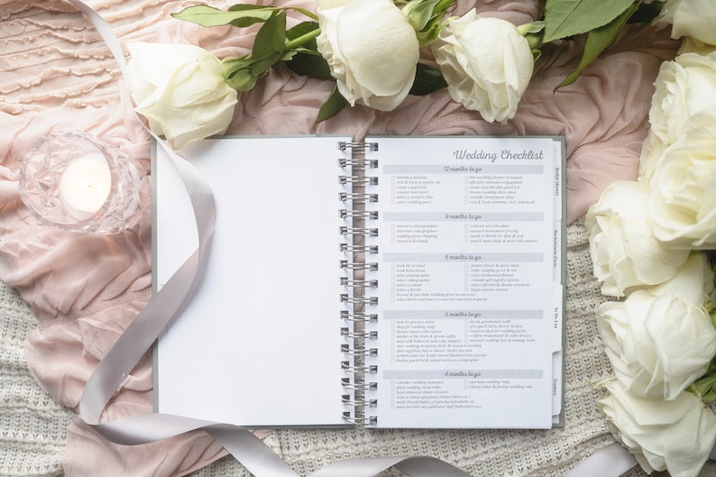 May include: An open wedding checklist planner with white roses and a candle on a pink and white fabric background. The planner has a checklist for 12 months, 8 months, 6 months, 5 months, and 4 months before the wedding.