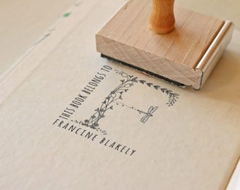 May include: A wooden stamp with a natural wood handle is on a page. The stamp has a floral design around the letter "F" and the text "THIS BOOK BELONGS TO FRANCINE BLAKELY". The stamp is pressed onto the page.