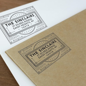 May include: Black ink address stamp on white and brown paper. The stamp reads "THE SINCLAIRS, 27 GILDED AVENUE, SAVOY HEIGHTS, NY 10019" in a decorative rectangular frame. The design is repeated on both papers.