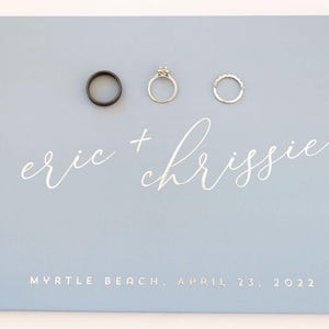 May include: A light blue wedding guest book with the names "eric + chrissie" written in silver script. The date "Myrtle Beach, April 23, 2022" is printed below the names. Three rings are displayed on the cover.