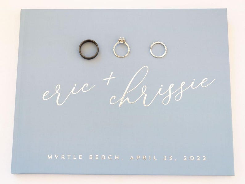 May include: A light blue wedding guest book with the names "eric + chrissie" written in silver script. The date "Myrtle Beach, April 23, 2022" is printed below the names. Three rings are displayed on the cover.