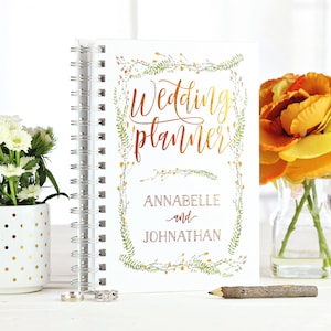 May include: A white spiral-bound wedding planner with gold lettering and floral accents. The cover reads "Wedding planner" with the names "ANNABELLE and JOHNATHAN" below. The planner is next to a vase of yellow flowers and a white pot with gold polka dots.