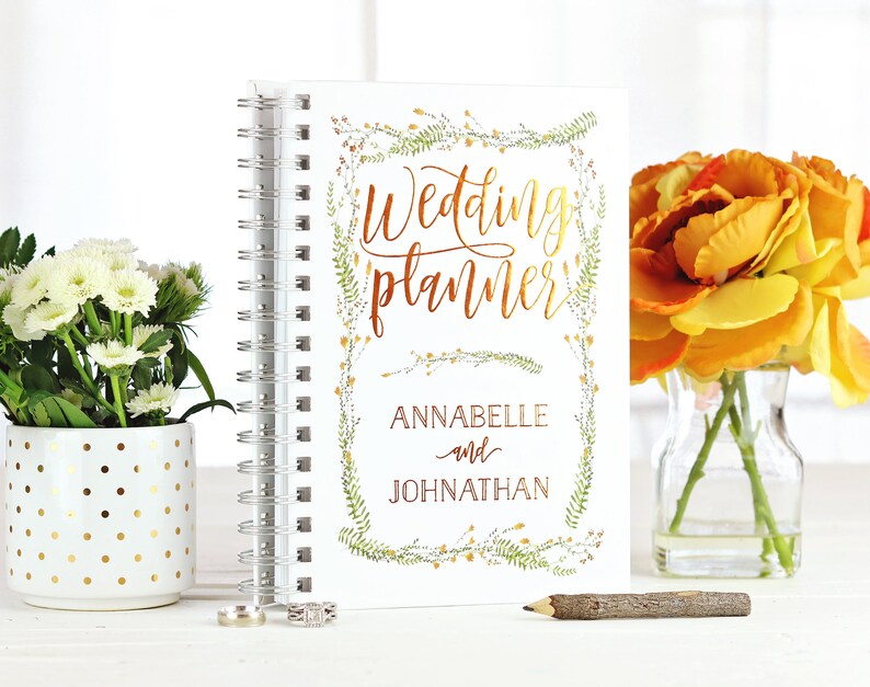 May include: A white spiral-bound wedding planner with gold lettering and floral accents. The cover reads "Wedding planner" with the names "ANNABELLE and JOHNATHAN" below. The planner is next to a vase of yellow flowers and a white pot with gold polka dots.