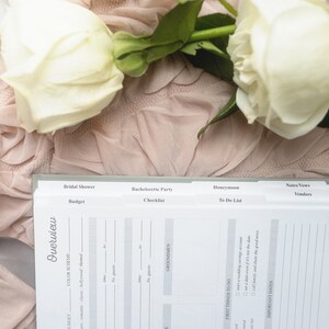 May include: A wedding planner notebook with labeled sections for Bridal Shower, Bachelorette Party, and Honeymoon. The notebook is open, revealing pages for budgeting, checklists, and to-do lists. White roses and a candle sit on a pink fabric.