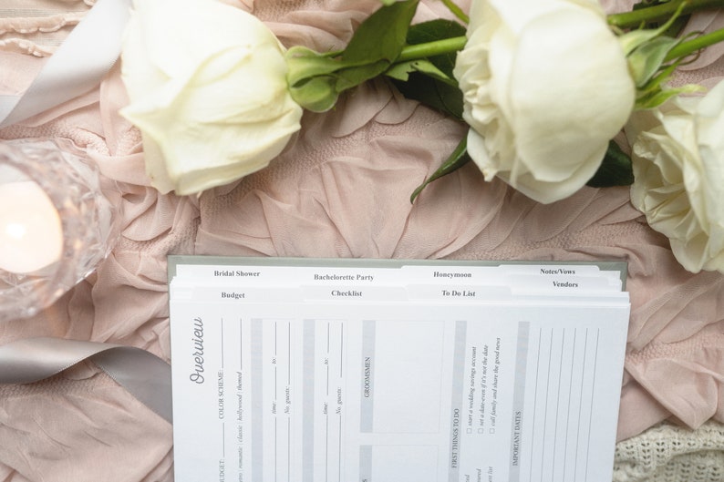 May include: A wedding planner notebook with labeled sections for Bridal Shower, Bachelorette Party, and Honeymoon. The notebook is open, revealing pages for budgeting, checklists, and to-do lists. White roses and a candle sit on a pink fabric.