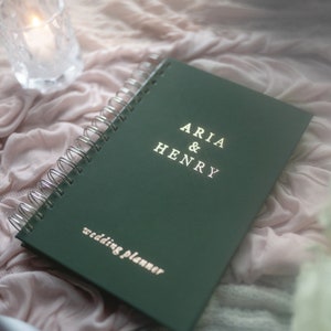 May include: A dark green wedding planner with silver spiral binding. The cover reads "ARIA & HENRY" above "wedding planner" in gold lettering. The planner rests on a pink fabric with a lit candle in a glass holder.