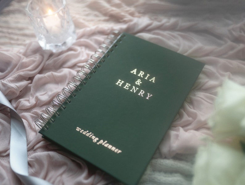 May include: A dark green wedding planner with silver spiral binding. The cover reads "ARIA & HENRY" above "wedding planner" in gold lettering. The planner rests on a pink fabric with a lit candle in a glass holder.