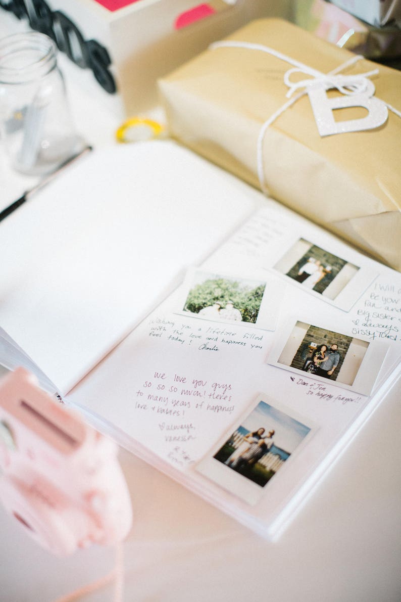 Może przedstawiać: Biały album ze zdjęciami z ręcznie napisanymi wiadomościami i zdjęciami Polaroid. Zdjęcia przedstawiają parę oraz ich przyjaci&oacute;ł i rodzinę. Album jest otwarty na stronie z wiadomością "We love you guys so so so much! There's to many years of happiness love + laughter!"
